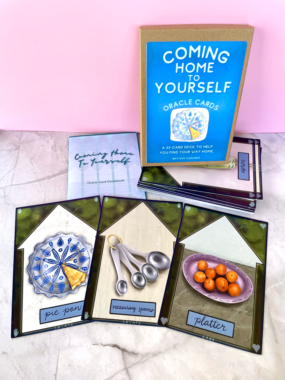 Coming Home to Yourself Oracle Card Deck created by Unruly Wit; photo of card samples laid out in front of the box on a marble countertop with a light pink backdrop.
