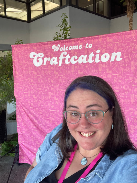 Brittany standing in front of a large pink backdrop that says “Welcome to Craftcation”.